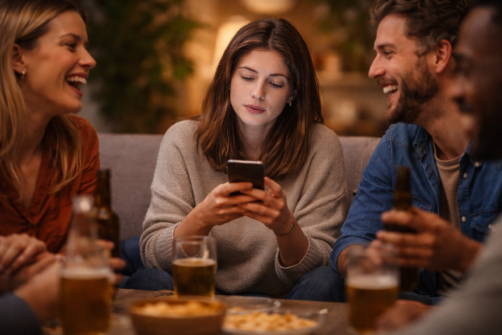 Frau schaut in einer Runde von Freunden auf ihr Smartphone, während die anderen miteinander lachen