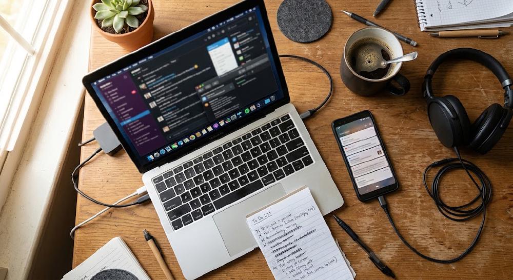 Schreibtisch von oben: Laptop mit vielen offenen Chat- und Mailfenstern, daneben ein Smartphone voller Benachrichtigungen, Kopfhörer, Kaffee und eine handschriftliche To-do-Liste